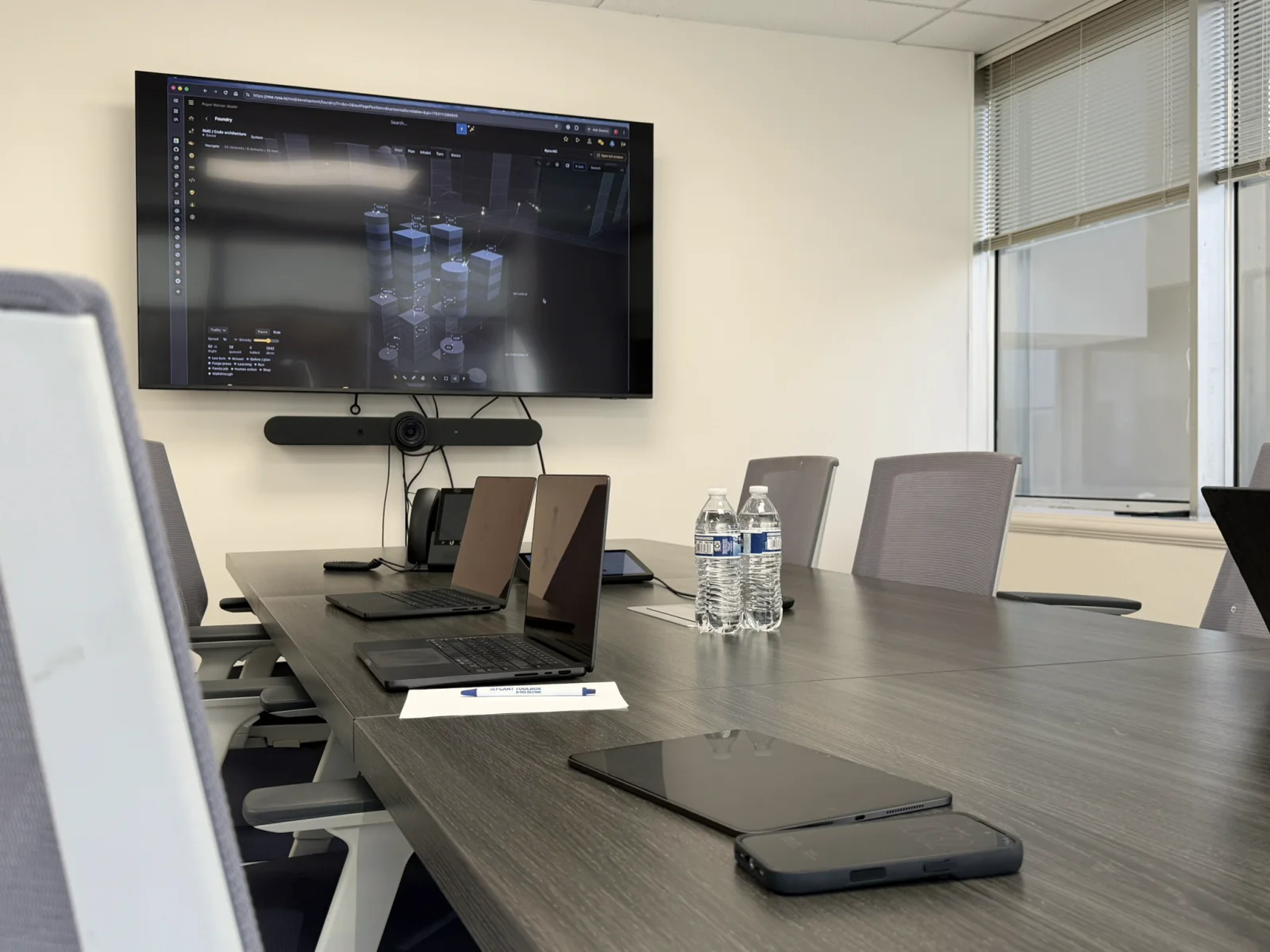 A conference room table set with laptops, a tablet and water, facing a wall screen with a video conferencing camera.